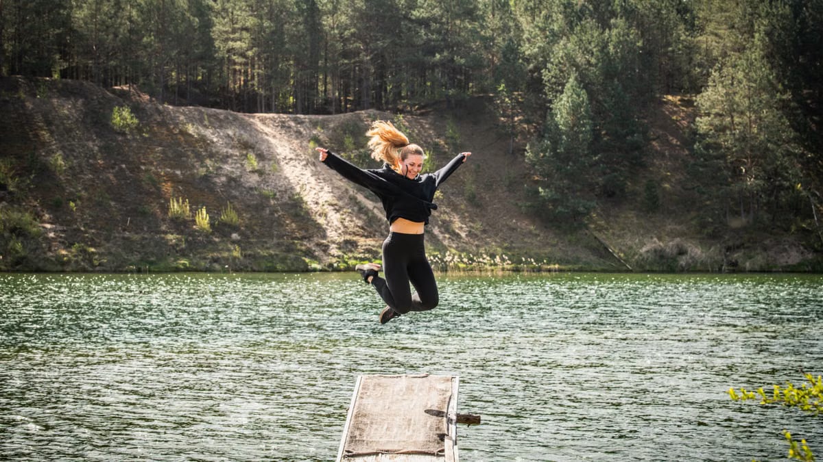 A woman leaps joyfully from a pier into a scenic forested lake in Lithuania, embracing adventure.